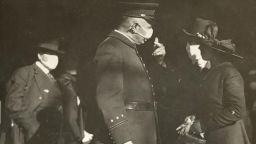 View of a policeman wearing a flu mask talking to a couple. The man is wearing a mask but woman is not, prompting a warning from the policeman. Others wearing masks can be seen in the background. Text written under photo: "Get one like this Right Away."