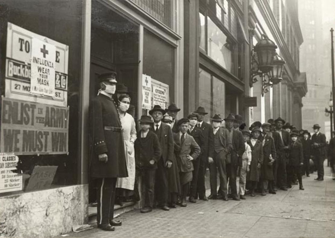 A line of people waiting for flu masksin 1918 in San Francisco. A sign on the window bearing the Red Cross logo reads: "Influenza. Wear Your Mask."