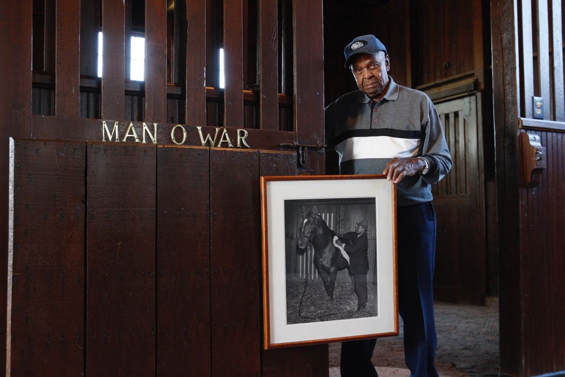 Tom Harbut, son of Man O' War's legendary groom Will Harbut, poses in 2010 with a vintage photograph of his father grooming the great sire by the stall where it was stabled.