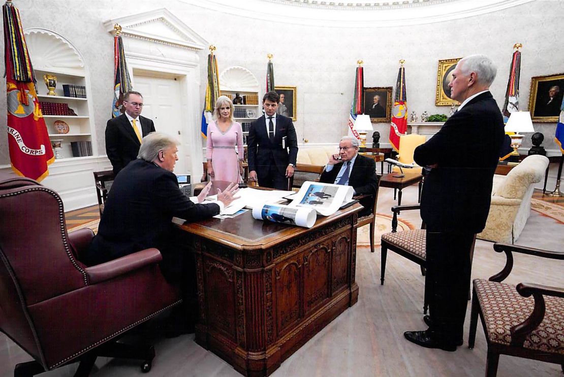 In this White House photo from December 2019 provided by Bob Woodward, President Donald Trump is seen speaking to Woodward in the Oval Office, surrounded by some aides and advisers, as well as Vice President Mike Pence. On Trump's desk is a large picture of Trump and North Korean leader Kim Jong Un.