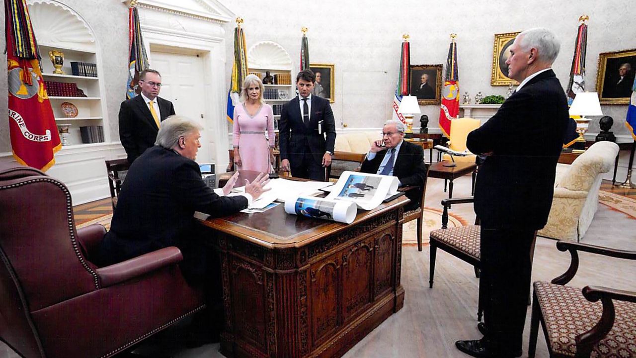 In this White House photo from December 2019 provided by Bob Woodward, President Donald Trump is seen speaking to Woodward in the Oval Office, surrounded by some aides and advisers, as well as Vice President Mike Pence. On Trump's desk is a large picture of Trump and North Korean leader Kim Jong Un.