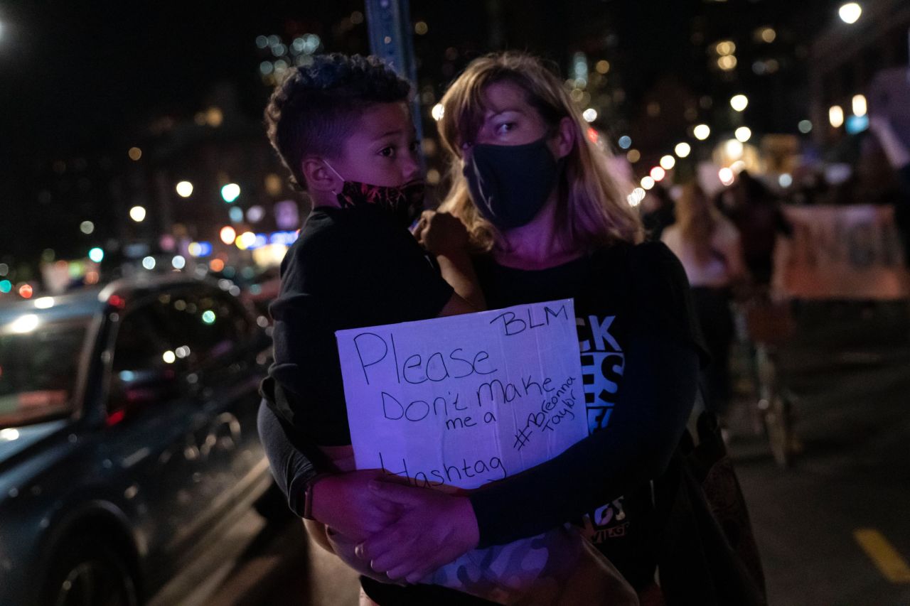 A woman carries a boy and a sign that reads, "Please don't make me a Breonna Taylor," as they watch protesters gather in Brooklyn, New York.