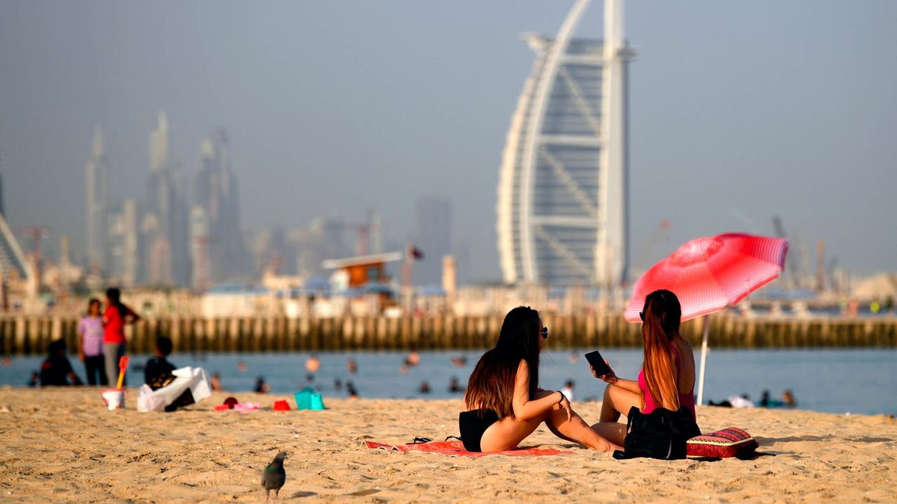 Woman sunbathers sit along a beach in the Gulf emirate of Dubai on July 24, 2020, while behind is seen the Burj al-Arab hotel. - After a painful four-month tourism shutdown that ended earlier in the month, Dubai is billing itself as a safe destination with the resources to ward off coronavirus. The emirate, which had 16.7 million visitors last year, had opened its doors to tourists despite global travel restrictions and the onset of the scorching Gulf summer in the hopes the sector will reboot before high season begins in the last quarter of 2020. (Photo by Karim SAHIB / AFP) (Photo by KARIM SAHIB/AFP via Getty Images)