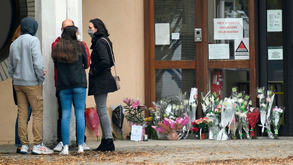 People stand next to flowers displayed at the entrance of the school in Conflans-Sainte-Honorine.