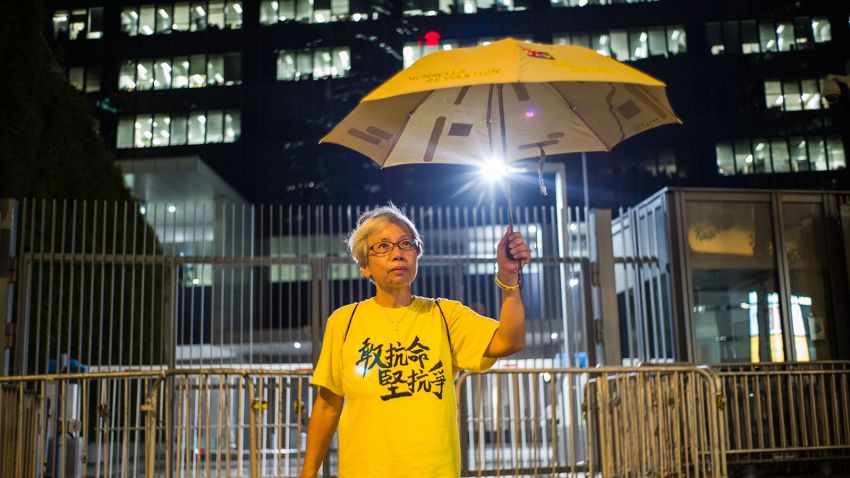 TOPSHOT - In this picture taken on September 27, 2016, retiree Alexandra Wong, 60, who lives in the neighbouring Chinese city of Shenzhen, poses in front of barricades in place for the second anniversary of the Umbrella Movement as she holds a yellow umbrella next to her art drawn on one of the government headquarter's driveways in Hong Kong, on the eve of the second anniversary of the Umbrella Movement. - The sprawling protest camps at the heart of Hong Kong's 2014 pro-democracy "Umbrella Movement" have long gone -- but artist Alexandra Wong is determined to keep the memory alive. Every week the 60-year-old returns to a spot on the driveway outside the city's government headquarters, creating political murals from brightly-coloured tape laid on the tarmac. (Photo by Anthony WALLACE / AFP) / To go with AFP Story:  "HONG KONG-CHINA-POLITICS-DEMOCRACY-ARTS", by Aaron Tam        (Photo credit should read ANTHONY WALLACE/AFP via Getty Images)