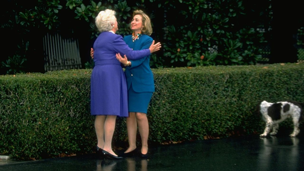 Hillary Rodham Clinton and first lady Barbara Bush exchange hugs as dog Millie sniffs hedge, on the grounds of the White House on November 19, 1992.