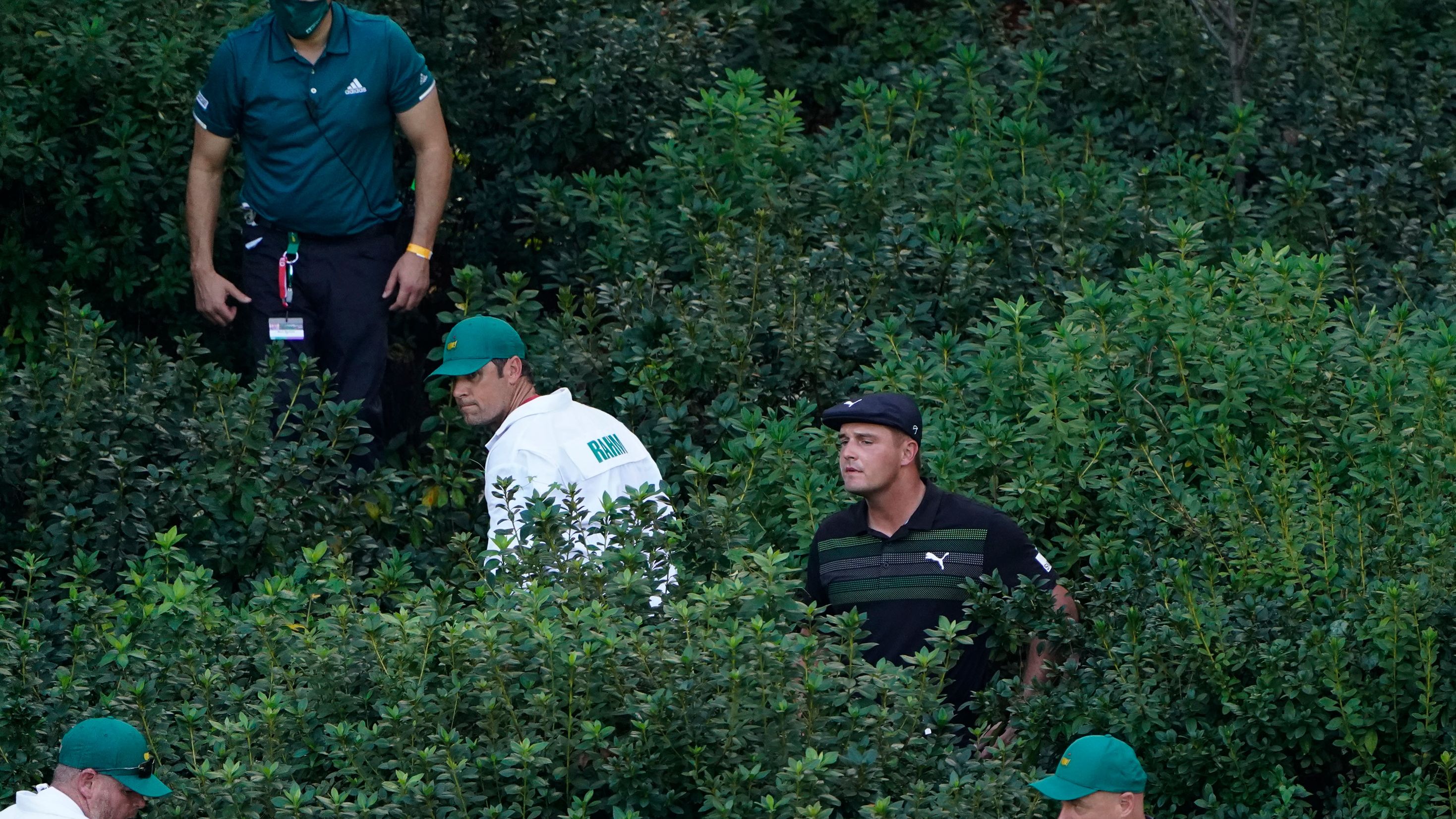 Bryson DeChambeau is aided by caddies and officials to look for his ball in the bushes on the 13th hole after a wayward shot.