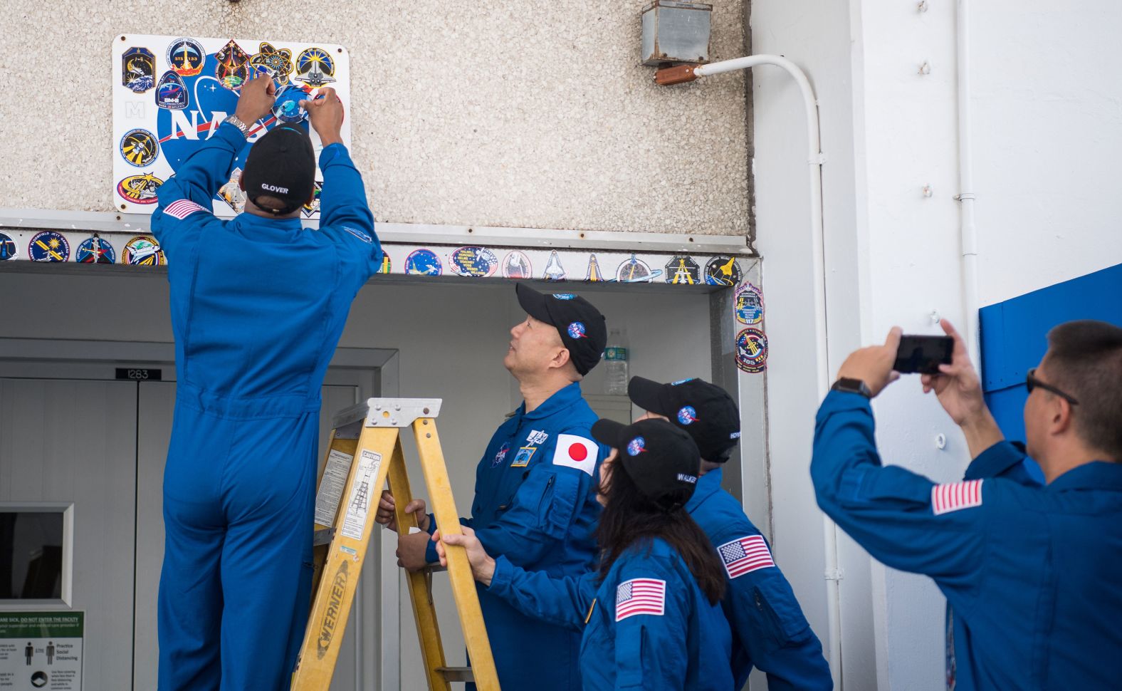 Glover places a Crew-1 mission sticker above the doorway to crew quarters at the Neil Armstrong Operations and Checkout Building as his fellow crewmates look on, November 12 at Kennedy Space Center.