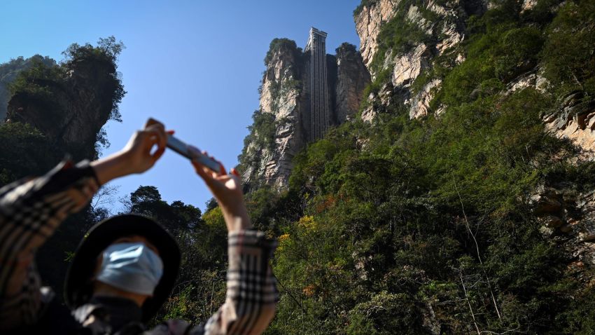 This picture taken on November 13, 2020 shows a tourist with a face mask using her mobil phone to take a picture at the entrance of the Bailong elevators in Zhangjiajie, China's Hunan province. - Towering more than 300 metres (1,000 feet) up the cliff face that inspired the landscape for the blockbuster movie "Avatar", the world's highest outdoor lift whisks brave tourists to breathtaking views. (Photo by WANG ZHAO / AFP) / TO GO WITH: China technology film tourism transport, by Ludovic EHRET (Photo by WANG ZHAO/AFP via Getty Images)