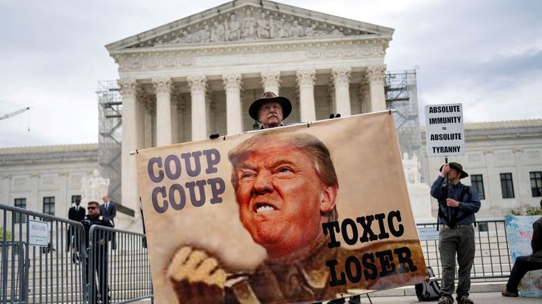 A White man with a white mustache and brown fedora hat stands outside the U.S. Supreme Court. He is holding a banner with Donald Trump's face on it, along with the words "Coup Coup Toxic Loser"
