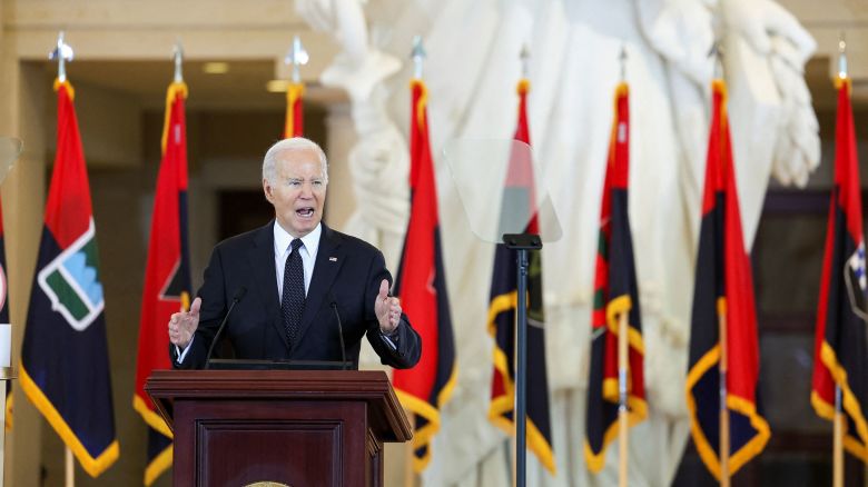 U.S. President Joe Biden speaks at a podium, in front of a row of red, navy and yellow flags. 