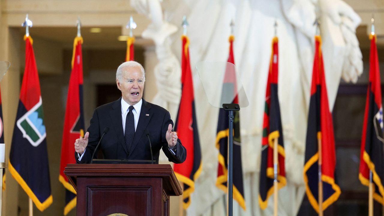 U.S. President Joe Biden speaks at a podium, in front of a row of red, navy and yellow flags. 