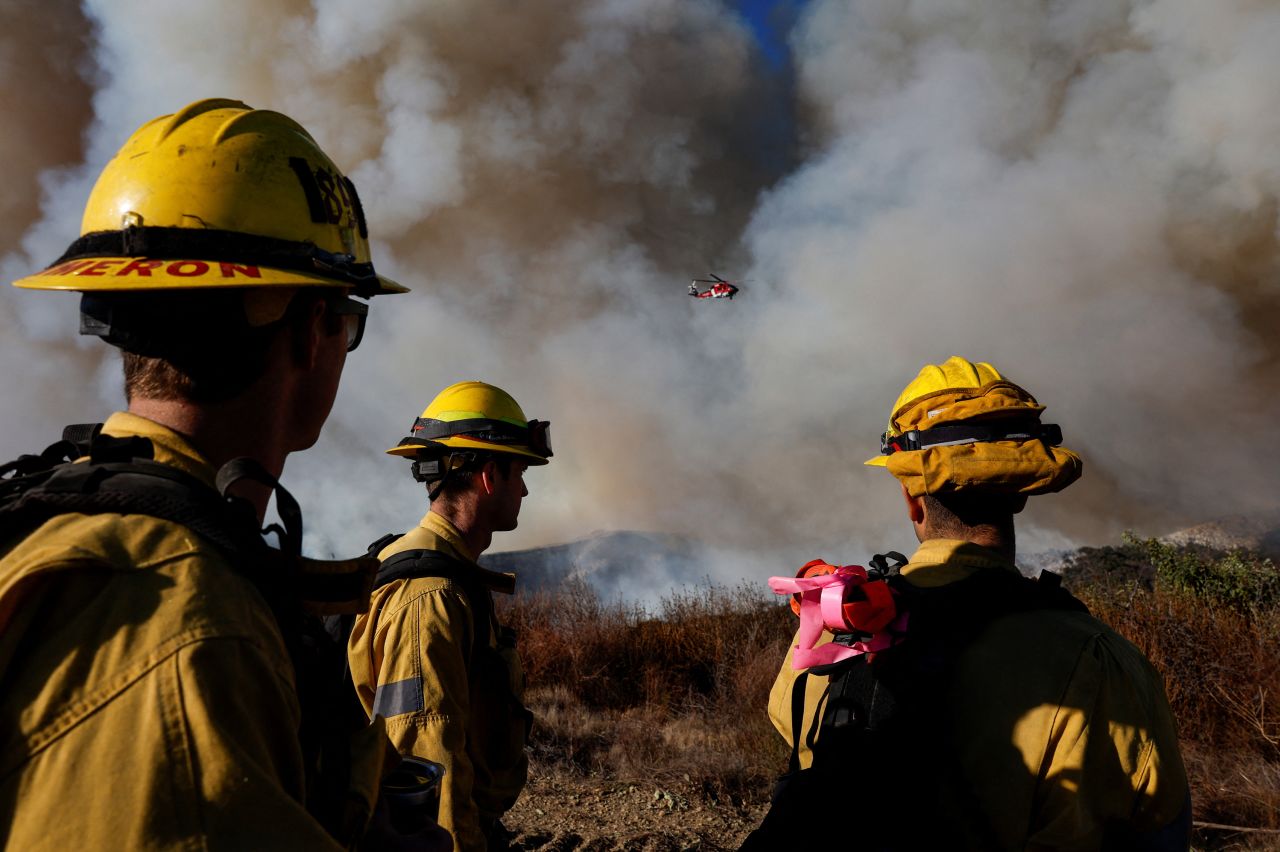 firefighters in yellow helmets and yellow helmets watch a large plume of smoke