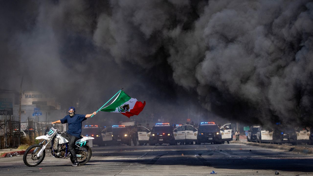 A man on a motorcycle waves a Mexican flag as smoke rises from a burning car on Atlantic Boulevard, during a standoff by protesters and law enforcement, following multiple detentions by Immigration and Customs Enforcement (ICE), in the Los Angeles County city of Paramount, California, U.S., June 7, 2025.