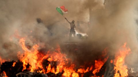 A man waves a Mexican flag as smoke and flames rise from a burning vehicle during a protest against federal immigration sweeps, near Los Angeles City Hall in downtown Los Angeles, California, U.S. June 8, 2025.