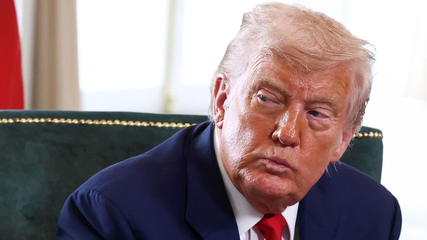 Donald Trump looks to the side during a meeting, wearing a navy blue suit and red tie, seated on a green velvet chair with gold trim.