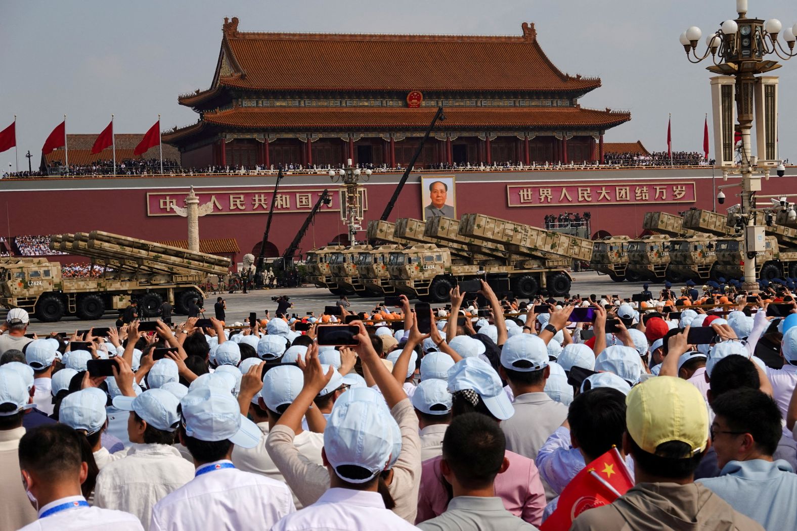 Spectators in caps photograph military vehicles as the parade passes by.