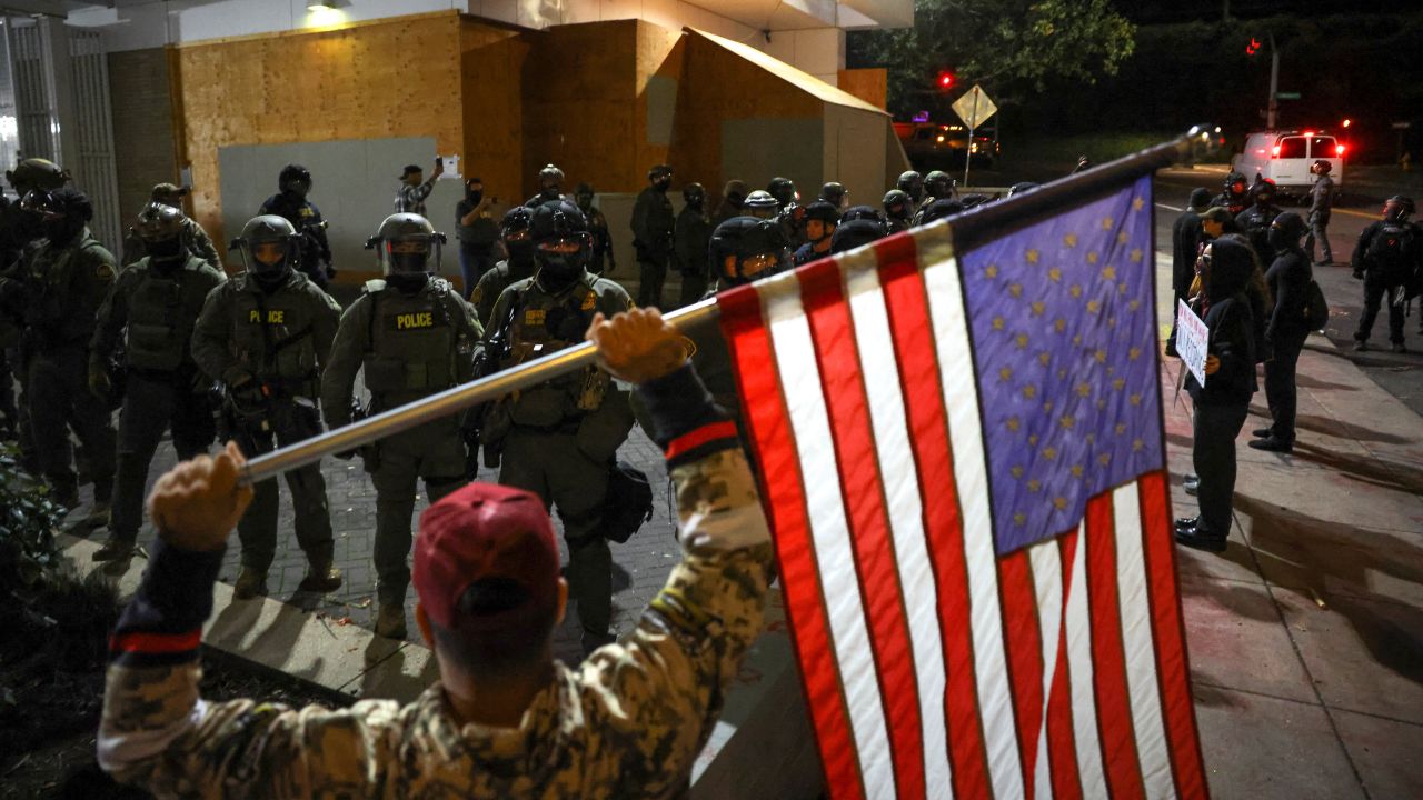 A man holds an American flag as law enforcement officers guard the enterance to ICE (Immigration and Customs Enforcement) headquarters in south Portland, Oregon, U.S., October 3, 2025. REUTERS/Carlos Barria