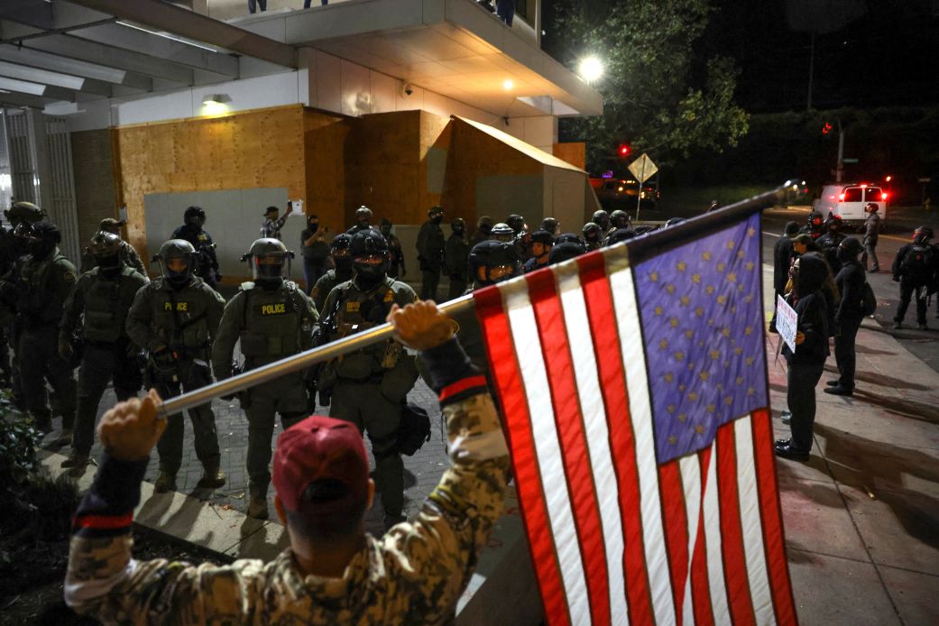 A man holds an American flag as law enforcement officers guard the enterance to ICE headquarters in south Portland, Oregon on Friday.