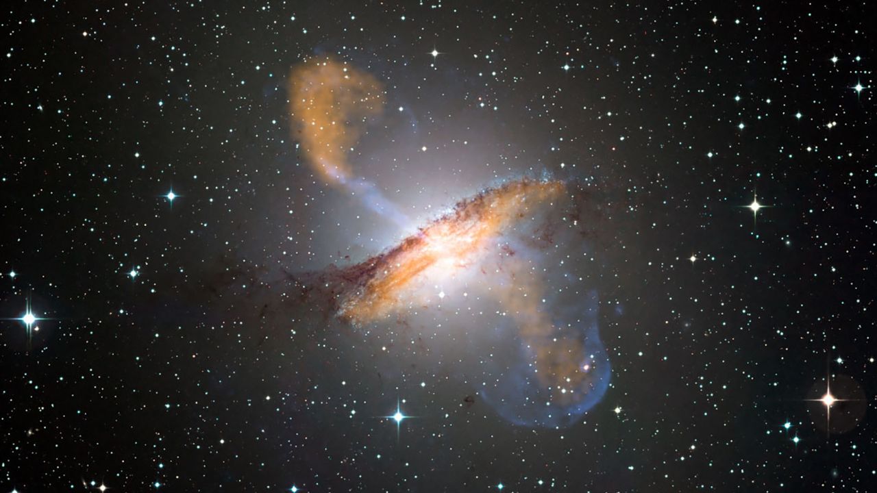 This composite image shows the Centaurus A galaxy in visible, microwave (orange) and X-ray (blue) light. Long jets expand out from the galaxy's central black hole on either side. 
