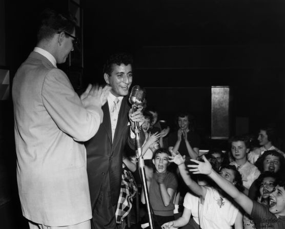 Bennett performs on a stage in Cleveland as local DJ Bill Randall applauds and young girls scream in the audience. Bennett had a string of hits in the early to mid-1950s, including chart-toppers "Because of You" and "Rags to Riches."
