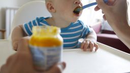 Baby taking a bite of food from a spoon with jar of baby food in foreground - stock photo
