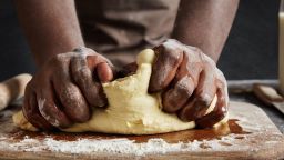 Unrecognizable male kneads dough on wooden counter, bakes delicious pastry, uses high quality flour. African male cook works in restaurant or at home kitchen, has dirty hands, prepares homemade cake