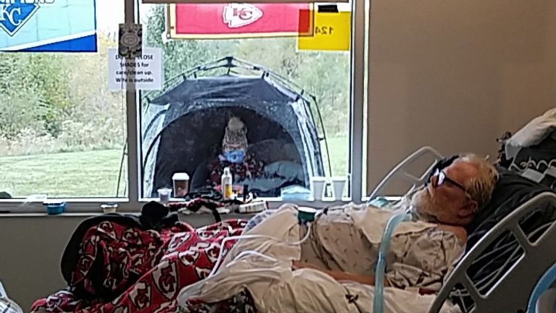 Roger Collins lies in his hospital bed while his wife, Billie, sits in a tent on the other side of the window.
