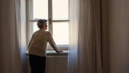 A portrait of a senior woman standing at home, looking out of a window.