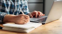 Male student writes a lecture in a notebook at home. He uses a laptop to watch a lecture.