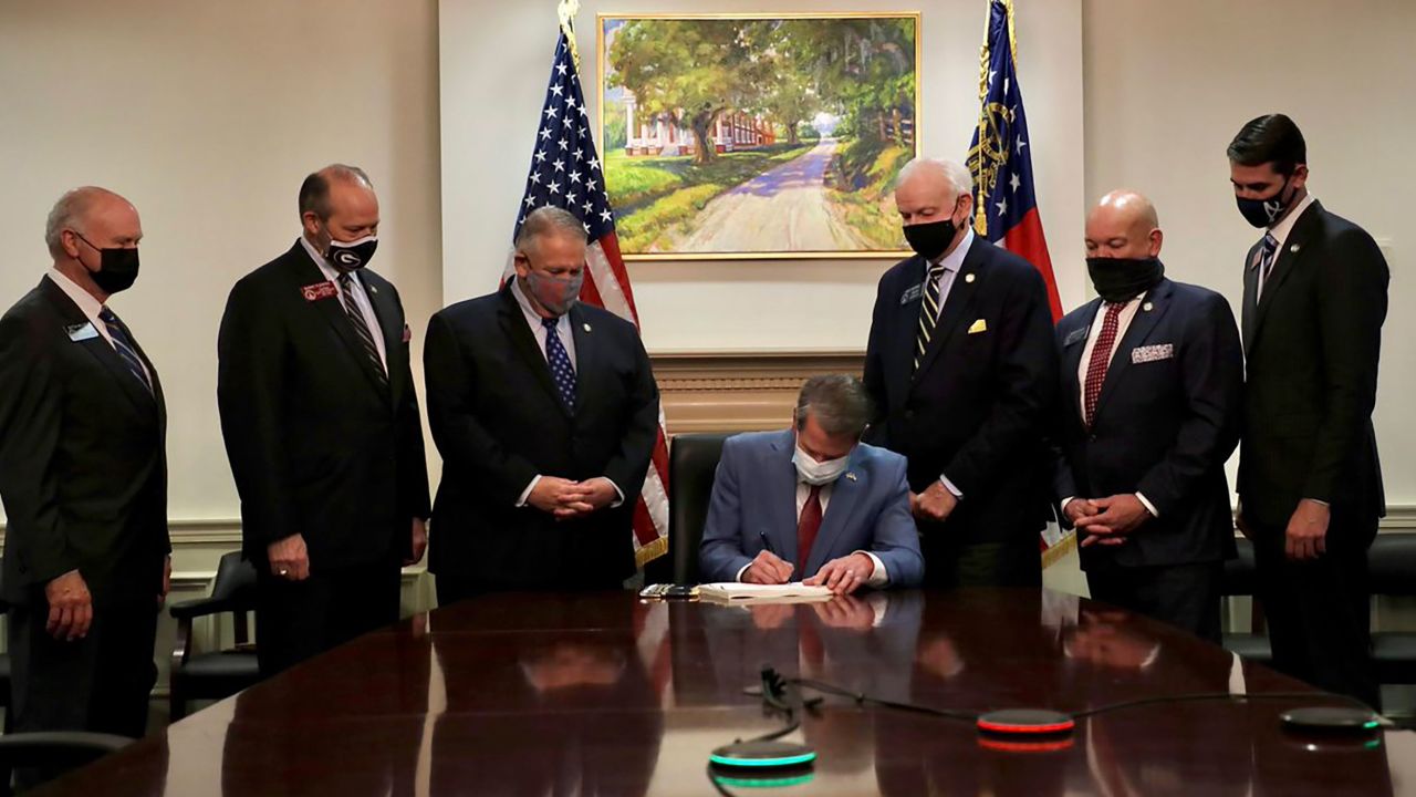 Georgia Gov. Brian Kemp signs SB202 as he sits at a table in a stately room, flanked by six men in suits and before a portrait of what seems to be a painting of an antebellum, plantation-styled home.



