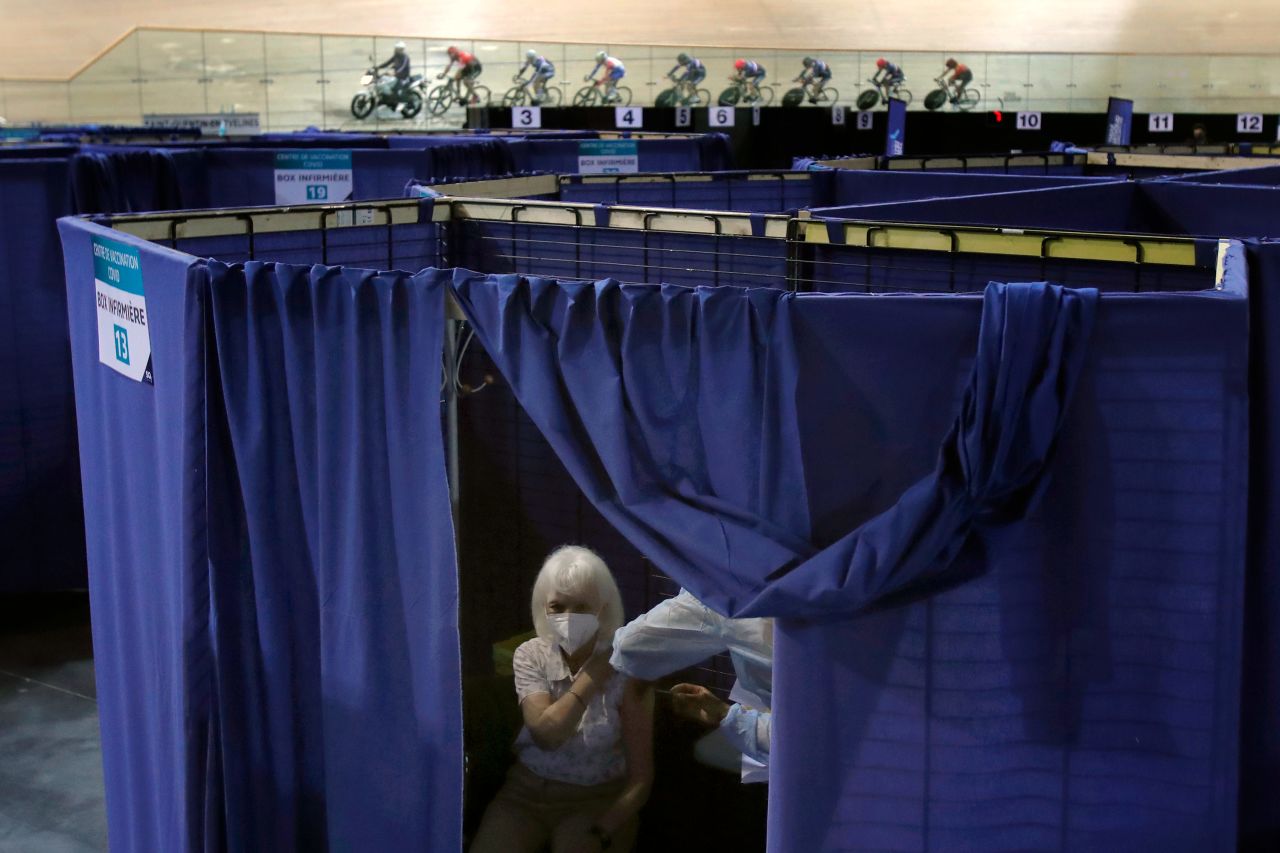 Members of the French cycling team are seen training in the background as a woman receives a Covid-19 vaccine at the National Velodrome in Saint-Quentin-en-Yvelines, France, on Monday, March 29. The velodrome has been turned into a mass vaccination center.