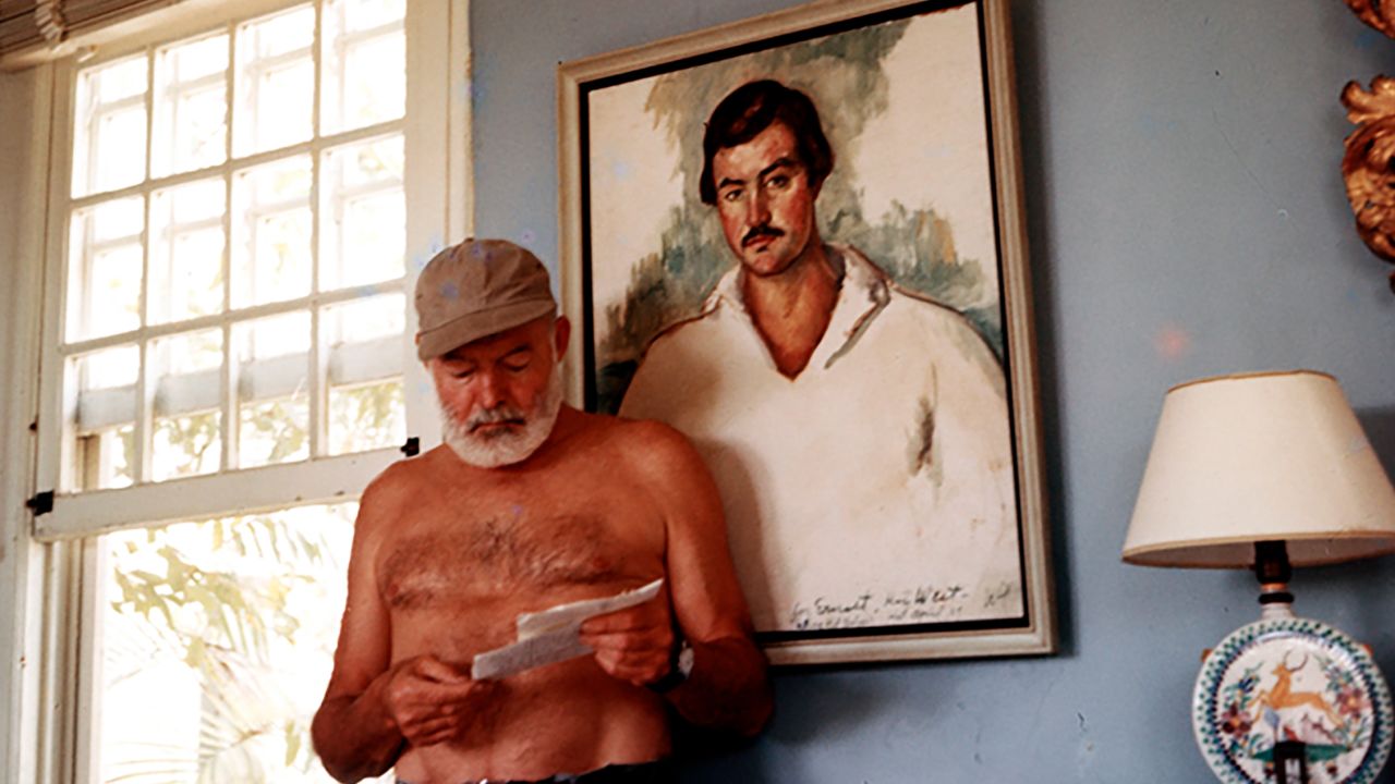 Ernest Hemingway at his home in Cuba, circa 1953, standing in front of a 1929 portrait of himself by Waldo Pierce.