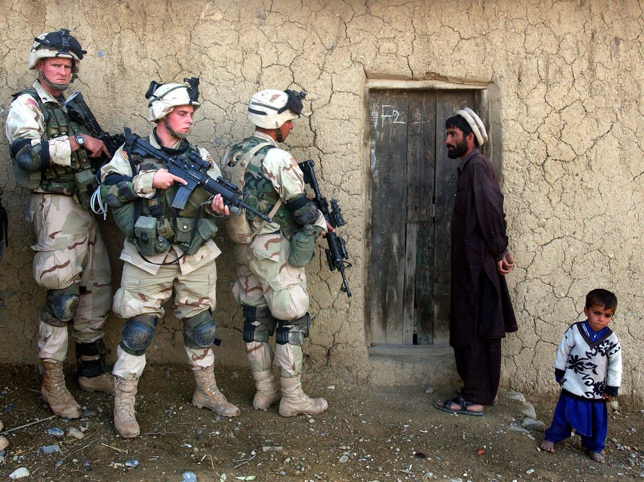 A man and his son watch US soldiers prepare to sweep their home in southeastern Afghanistan in November 2002.
