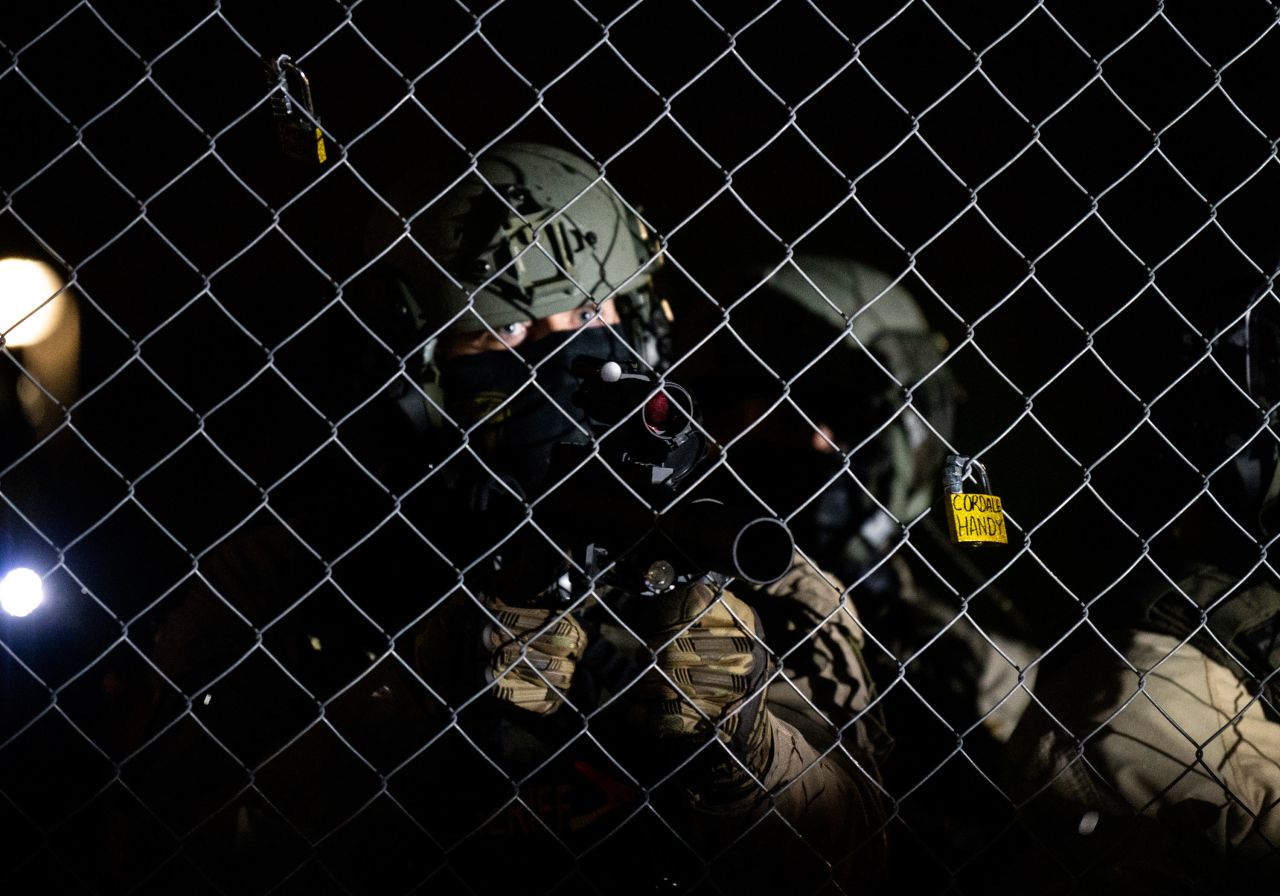 A law enforcement officer points a projectile weapon through fencing outside the police headquarters.