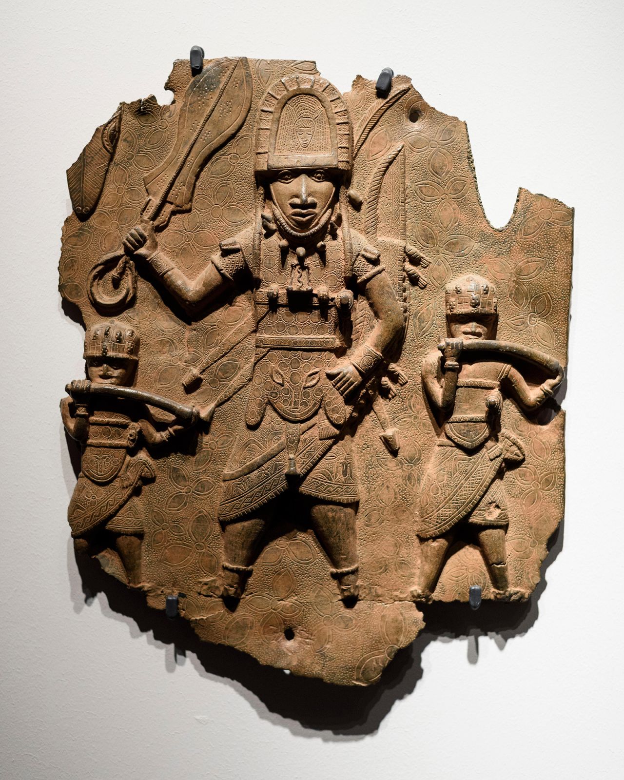 A Benin Bronze, pictured in Berlin, depicts a high-ranking dignitary with sword and rectangular bell accompanied by two hornblowers, brass plaque.