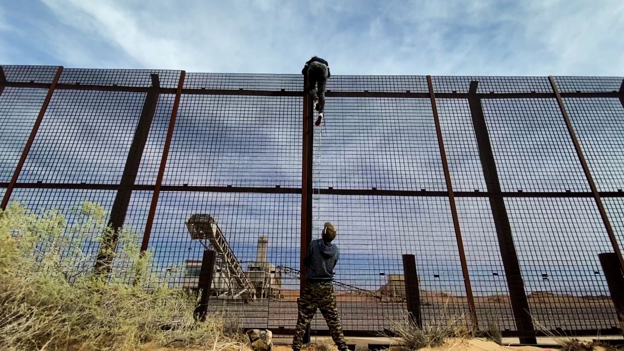 A migrant from Ecuador seen climbing over the US-Mexico border wall fence as a smuggler steadies the ladder below.