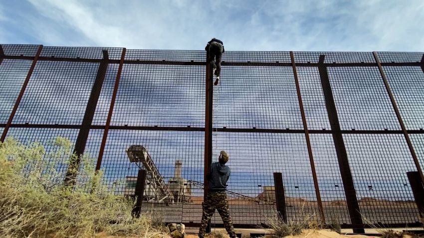 A 20 year-old female migrant from Ecuador is seen climbing over the US-Mexico border wall fence as the human smuggler that brought her to the wall steadies the ladder below.