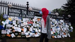 A woman passes a fence outside Brooklyn's Green-Wood Cemetery adorned with tributes to victims of COVID-19, Thursday, May 28, 2020, in New York. The memorial is part of the Naming the Lost project which attempts to humanize the victims who are often just listed as statistics. The wall features banners that say "Naming the Lost" in six languages: English, Spanish, Mandarin, Arabic, Yiddish and Bengali. (AP Photo/Mark Lennihan)