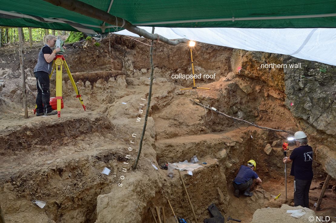 The decorated bone was found in the former entrance to the cave about 1 meter  behind the person holding the staff in the right of the image. 