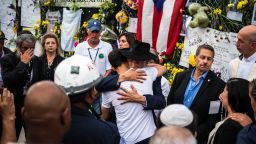 A member of the Miami-Dade Fire Rescue hugs family member and friends of the victims at the "Surfside Wall of Hope & Memorial" near the site where a building collapsed in Surfside, Florida, north of Miami Beach on July 7, 2021. - Florida rescuers have made the "extremely difficult decision" to end their search for survivors in the rubble of an apartment building which partially collapsed nearly two weeks ago, Miami-Dade county mayor Daniella Levine Cava said July 7. "It is with deep profound sadness ... that we made the extremely difficult decision to transition from operation search and rescue to recovery," Levine Cava told reporters in Surfside, near Miami, adding that the transition would formally take place at midnight. (Photo by CHANDAN KHANNA / AFP) (Photo by CHANDAN KHANNA/AFP via Getty Images)