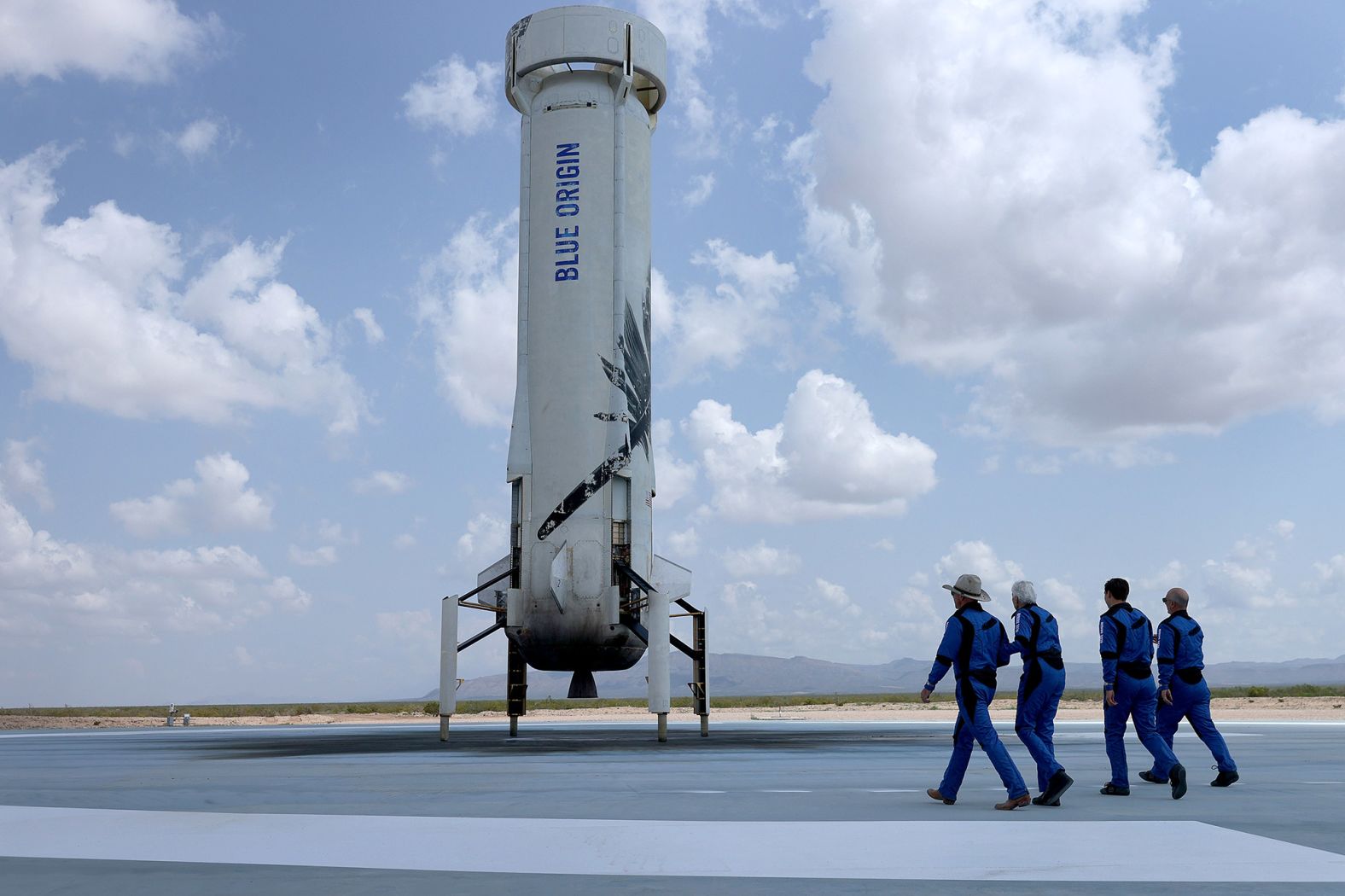 The crew walks toward the reusable rocket booster to pose for a picture after their flight. The booster launched the capsule and then returned to the ground and landed upright.
