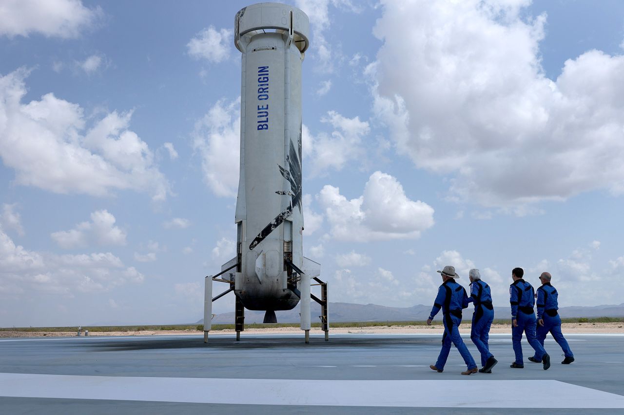 The crew walks toward the reusable rocket booster to pose for a picture after their flight. The booster launched the capsule and then returned to the ground and landed upright.