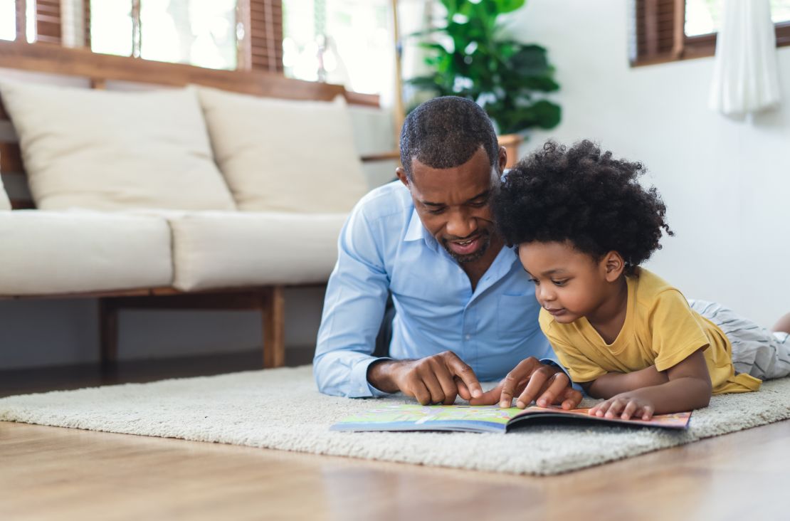 A father and his son read a book while lying on the floor at home.