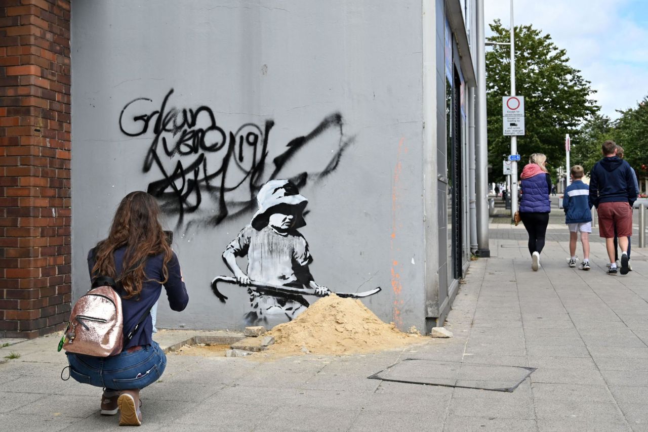 A woman takes a photograph of a stensil of a child digging in the sand, the subject of a graffiti artwork bearing the hallmarks of street artist Banksy on a wall in Lowestoft on the East Coast of England on August 8, 2021.