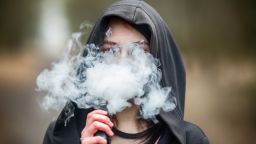 Вейп тийнейджър. Млада красива кавказка брюнетка, която пуши електронна цигара на улицата през пролетта. Смъртоносно лош навик. class=
