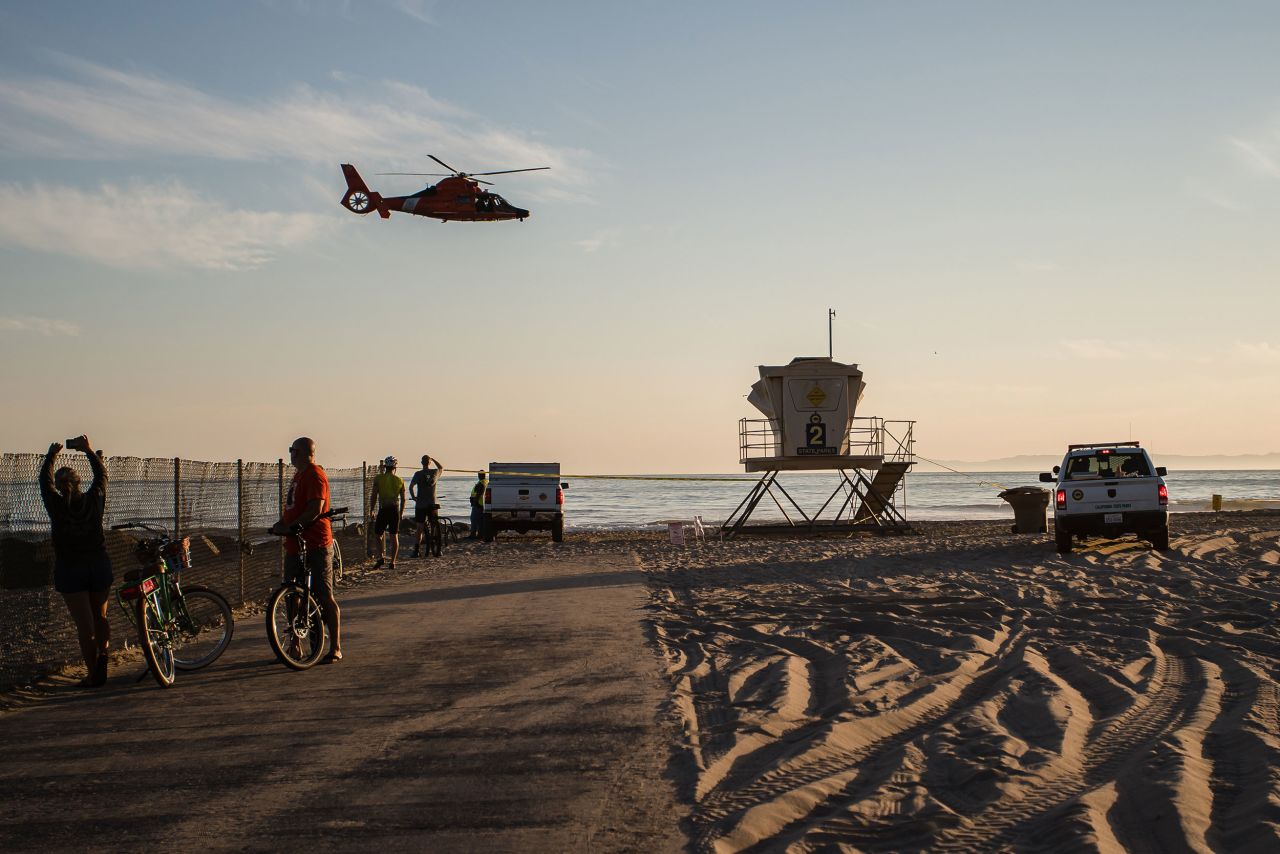A helicopter flies over bystanders in Huntington Beach. The federal Bureau of Safety and Environmental Enforcement is assisting in the Coast Guard-led response to the oil spill, the agency told CNN.