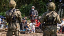 Polish (foreground) and Belarusian (background) border guards stand next to migrants believed to be from Afghanistan sitting on the ground in the small village of Usnarz Gorny near Bialystok, northeastern Poland, located close to the border with Belarus, on August 20, 2021. - The fate of a group of 32 bedraggled migrants stranded at a makeshift encampment on the border between Belarus and Poland for nearly two weeks has sparked a heated debate in Poland. (Photo by Wojtek RADWANSKI / AFP) (Photo by WOJTEK RADWANSKI/AFP via Getty Images)