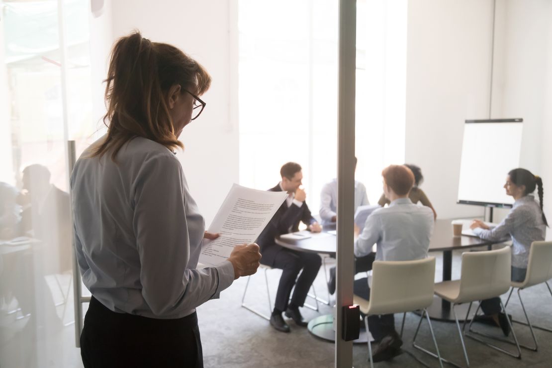 A businesswoman stands at an office door, feeling nervous before her presentation for a meeting. 