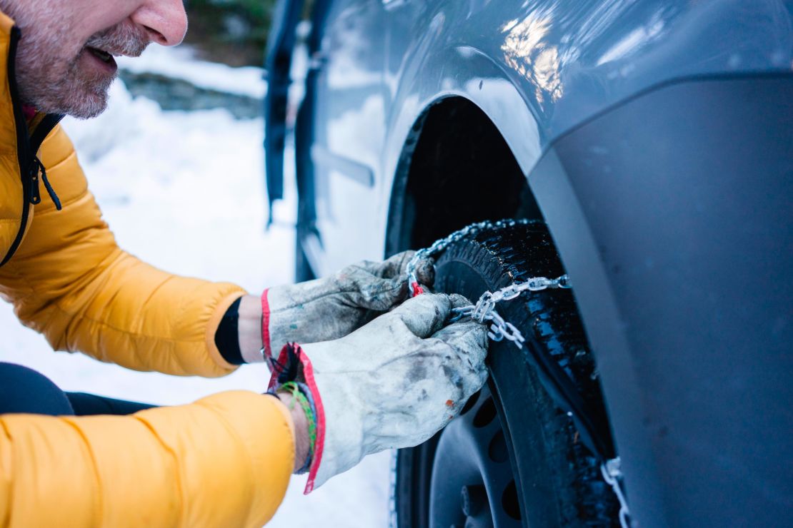 A person puts snow chains on a tire while parked in snowy conditions. 
